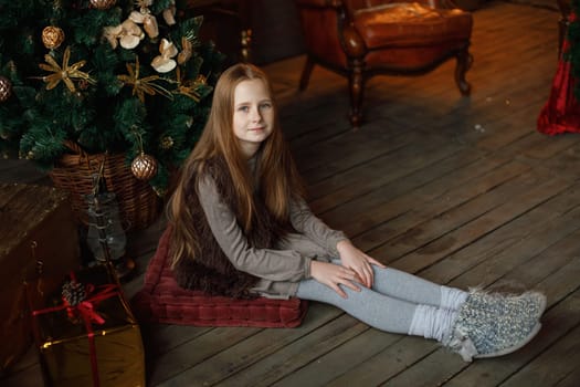 A young girl with long hair sits on a cushion on a wooden floor, surrounded by a festive Christmas tree adorned with ornaments. She wears a cozy outfit with a fur vest and knitted socks, creating a warm holiday atmosphere.