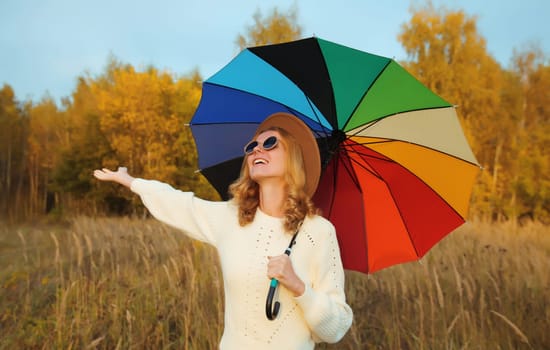 Happy young woman with colorful umbrella in autumn park raising her hands up, enjoying warm weather, wearing sweater, fall season background