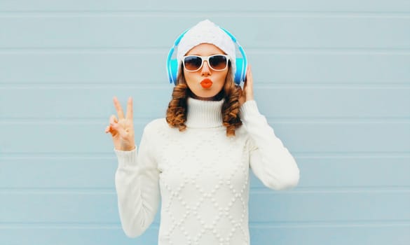 Fashion woman in headphones listens to music wear glasses, hat, sweater over blue background