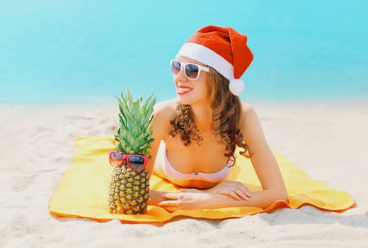 Christmas portrait young smiling woman in red santa hat and pineapple lying on beach on sea