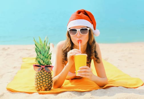 Christmas portrait woman in red santa hat with pineapple drinking juice lying on beach on sea