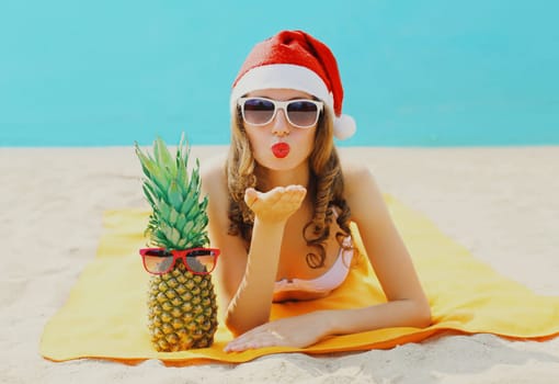 Christmas portrait woman in red santa hat with pineapple lying on beach blowing kiss on sea