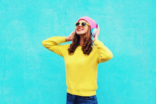 Happy young woman listening to music with headphones in colorful yellow sweater on blue background