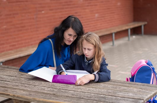 loving mother watching her daughter doing homework on her own at wooden table in children's playground, education concept, back to school, girl in beautiful blue uniform,