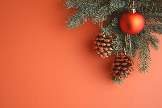 The image showcases a beautiful arrangement of Christmas decorations. A branch of evergreen holds three ornaments—a bright red ball and two glittering pinecones—against a warm orange backdrop.