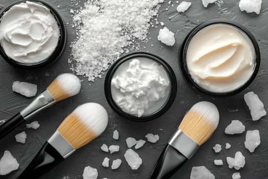 Skincare creams in black containers are displayed alongside soft brushes on a textured gray surface, accented by scattered salt crystals, creating a soothing arrangement.