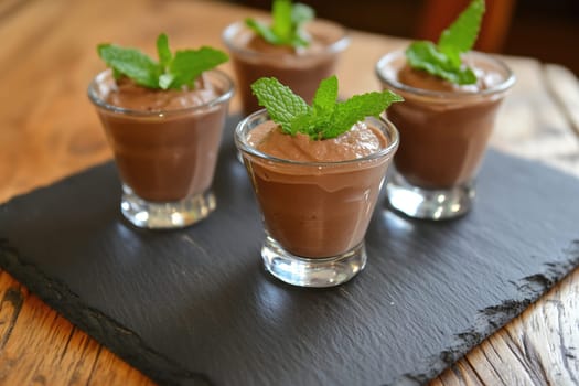 Five small glass cups filled with creamy chocolate mousse are beautifully garnished with fresh mint leaves, elegantly arranged on a dark slate platter.