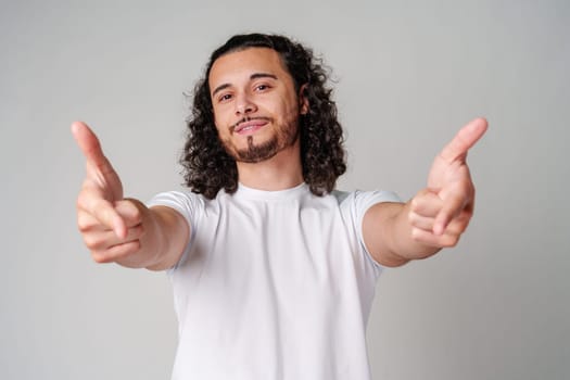 A smiling young man with curly hair playfully pointing directly at the viewer in a light-colored setting close up