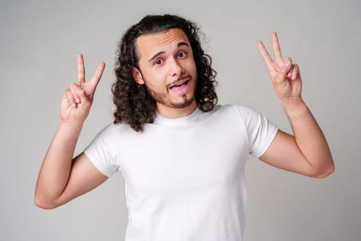 Young man with curly hair playfully posing with peace signs against a neutral background close up