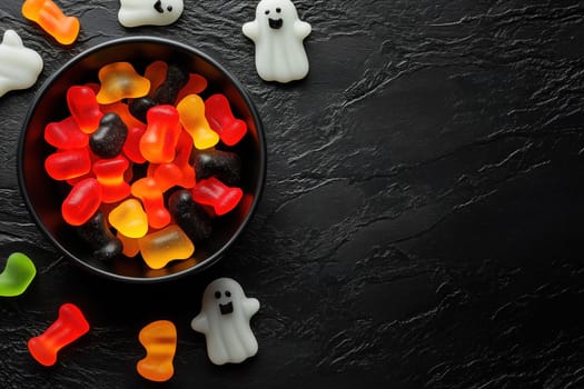 Colorful gummy candies, including ghost and bear shapes, are arranged in a black bowl on a textured black surface, perfect for Halloween treats or festive celebrations.