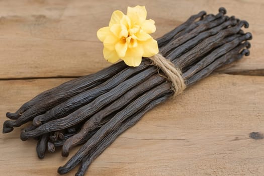 A bundle of dark vanilla bean pods is secured with twine, complemented by a delicate yellow flower, all resting on a wooden surface. The natural light enhances the rich textures.
