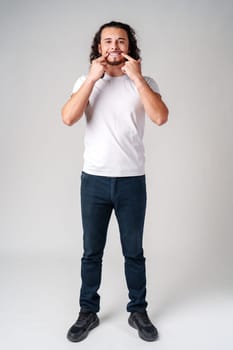 Young man in a white t-shirt playfully smiles while pointing to his teeth against a plain gray background in a studio setting