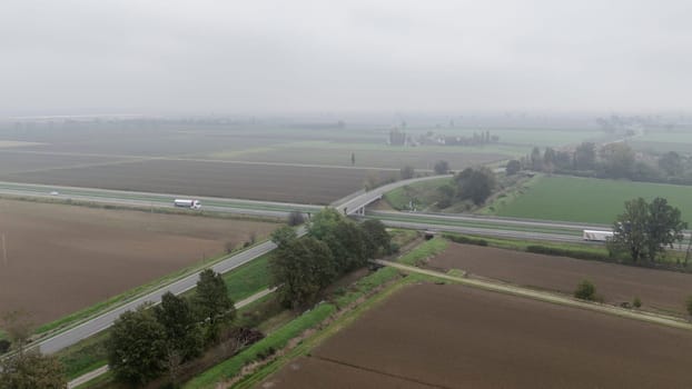 Piacenza, Italy October 15th 2024 Aerial view of a highway crossing the countryside on a foggy day, with trucks driving on the asphalt