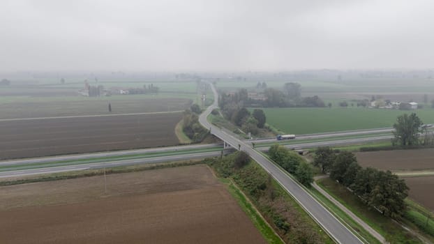 Piacenza, Italy October 15th 2024 An aerial view of a highway crossing over a bridge above a two lane road surrounded by farmland on a foggy day