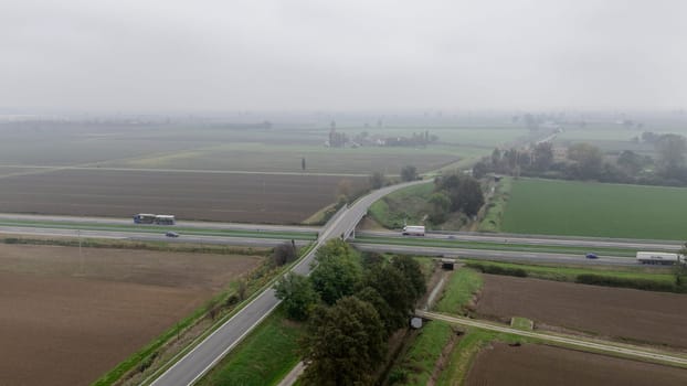 Piacenza, Italy October 15th 2024 Aerial view of a highway with cars and trucks passing over a countryside road on a foggy day