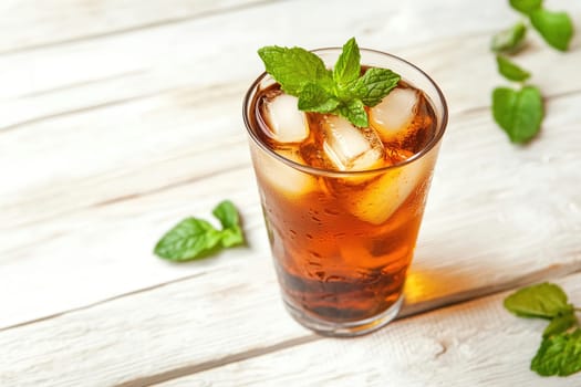 A tall glass filled with cold iced beverage and ice cubes is garnished with fresh mint leaves, placed on a rustic wooden table in the afternoon light.