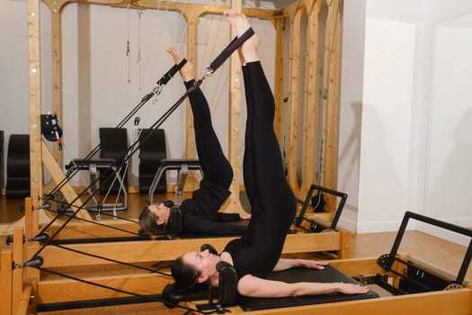 Two women in black sportswear are doing pilates on a reformer machine in the gym.