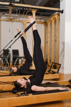 Two women in black sportswear are doing pilates on a reformer machine in the gym.
