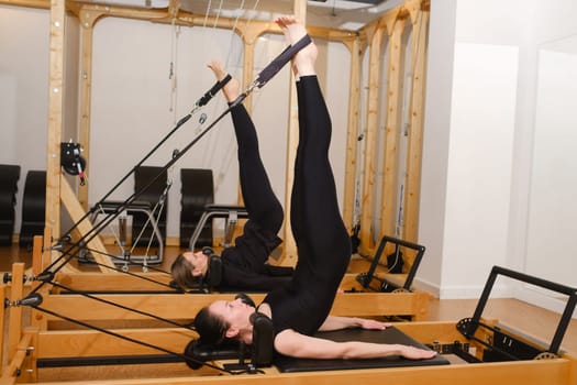 Two women in black sportswear are doing pilates on a reformer machine in the gym.