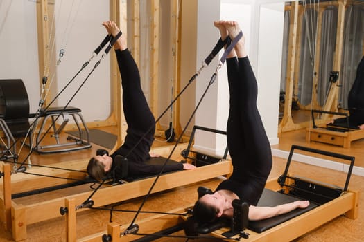 Two women in black sportswear are doing pilates on a reformer machine in the gym.