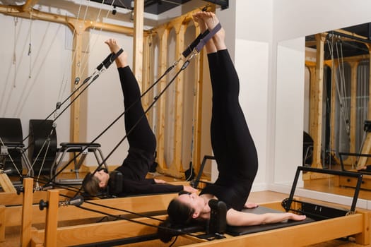 Two women in black sportswear are doing pilates on a reformer machine in the gym.