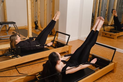 Two women in black sportswear are doing pilates on a reformer machine in the gym.