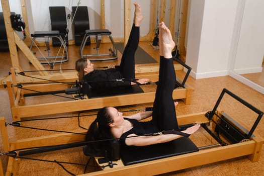Two women in black sportswear are doing pilates on a reformer machine in the gym.