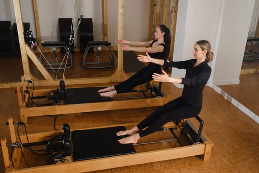 Two women sitting on a reformer exercise machine perform an exercise in the gym.