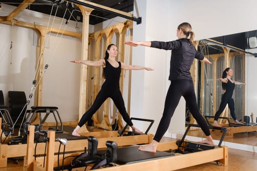 Two women standing on a reformer exercise machine perform an exercise in the gym.