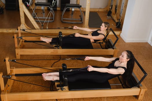 Two women in black sportswear are doing pilates on a reformer machine in the gym.