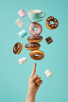 Hand holding a cup and saucer with floating donuts and marshmallows above it on a blue background. This whimsical image is perfect for advertising or social media