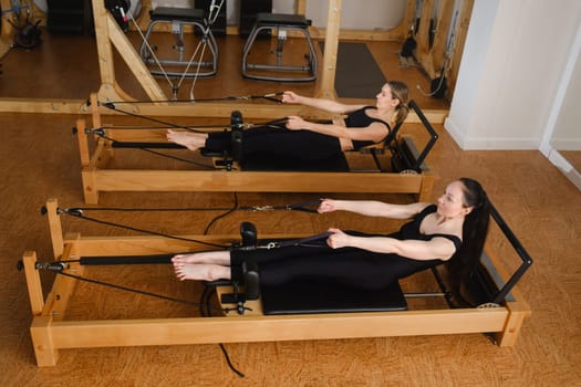 Two women in black sportswear are doing pilates on a reformer machine in the gym.