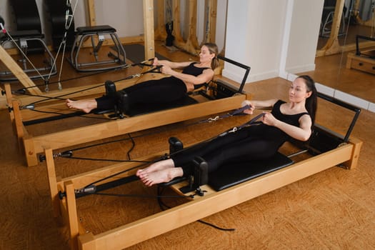 Two women in black sportswear are doing pilates on a reformer machine in the gym.