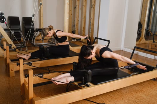 Two women in black sportswear are doing pilates on a reformer machine in the gym.