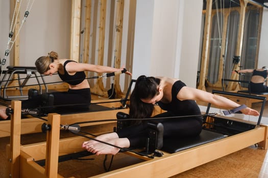 Two women in black sportswear are doing pilates on a reformer machine in the gym.