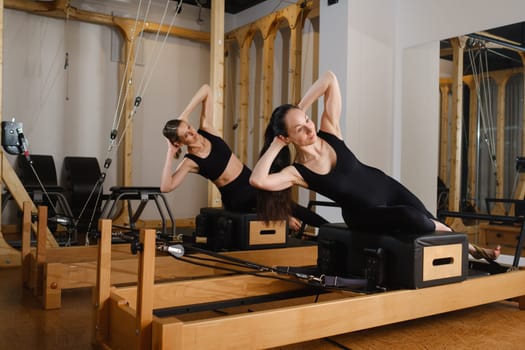 Two women in black sportswear are doing pilates on a reformer machine in the gym.