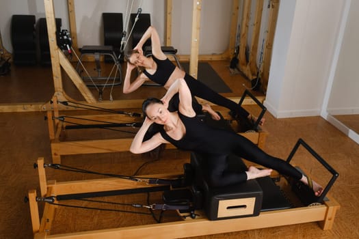 Two women in black sportswear are doing pilates on a reformer machine in the gym.
