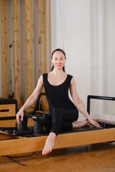 A young girl is doing Pilates on a reformer in the room. Beautiful slim woman in black clothes performs exercises to strengthen the spine and back muscles.