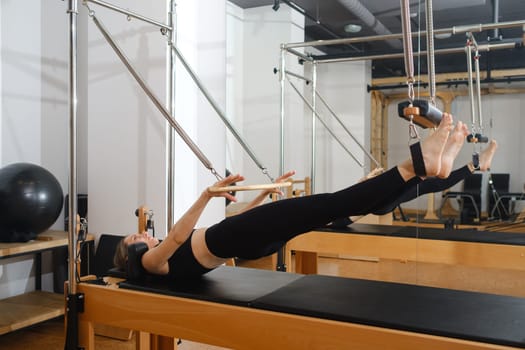Young girl doing Pilates on trapeze table in the gym. Beautiful slim woman in black clothes performs exercises to strengthen the spine and back muscles.