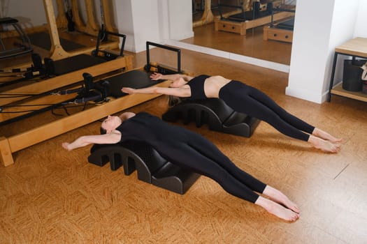 Two women are practicing physical fitness exercises on a Pilates machine in a room with hardwood flooring. Their movements showcase a beautiful balance of art and exercise