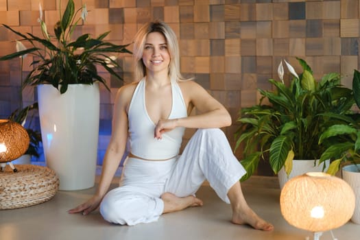 A young female yoga instructor in white athletic wear sits on the exercise floor in her studio before class.