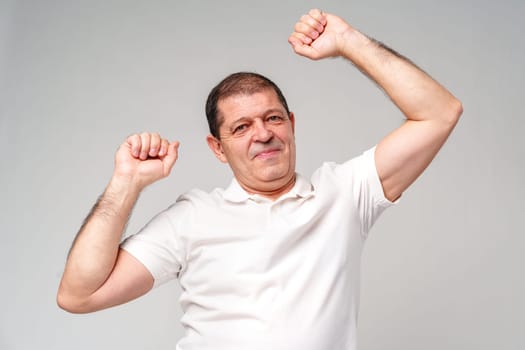 Happy older man showing off his strong biceps while standing against a light gray background close up