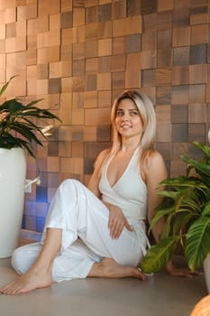 A young female yoga instructor in white athletic wear sits on the exercise floor in her studio before class.