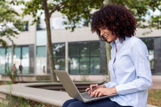 focused happy female entrepreneur with curly hair sitting on stone bench in park and working remotely on laptop