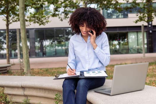smiling female entrepreneur sitting on stone bench in park and working remotely while speaking on smartphone and taking notes in documents