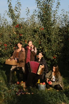 Four fashionable country girls at the apple harvest in the orchard. Happy girls playing accordion after apple harvest in an autumn orchard.