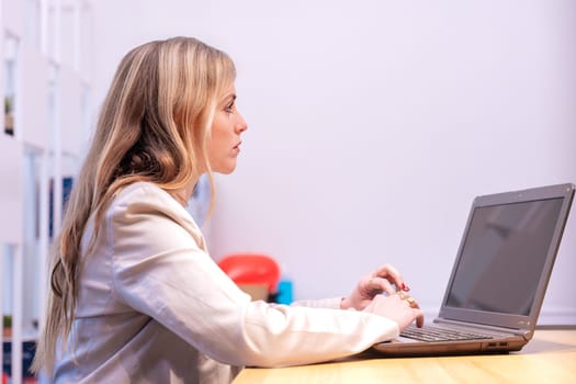 professional woman wearing a light-colored blazer sits at a wooden desk using a laptop in a brightly lit office. The setting includes subtle background details like colorful objects.