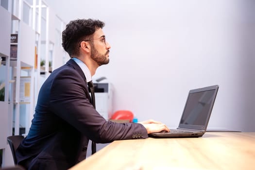 professional man wearing a light-colored blazer sits at a wooden desk using a laptop in a brightly lit office. The setting includes subtle background details like colorful objects.