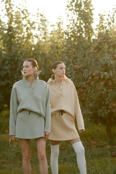 Two models in stylish tracksuits pose in apple orchard at sunset . The model is wearing a light-colored tracksuit and skirt.