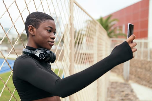 young ethnic female with short hair wearing wireless headset taking self portrait on mobile phone against grid fence